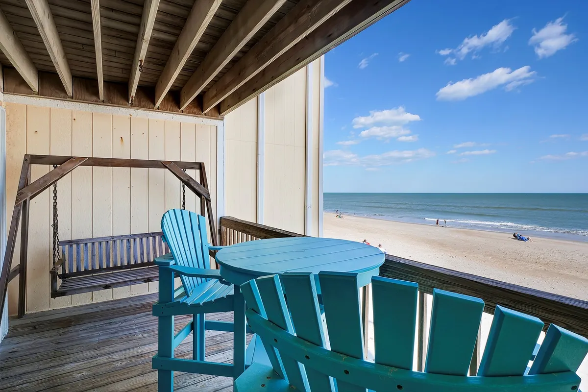 A tranquil exterior scene featuring a swing bench and blue chairs on a balcony overlooking the ocean. The golden light suggests sunset, and the gentle waves can be seen in the background, inviting relaxation and peaceful conversation.