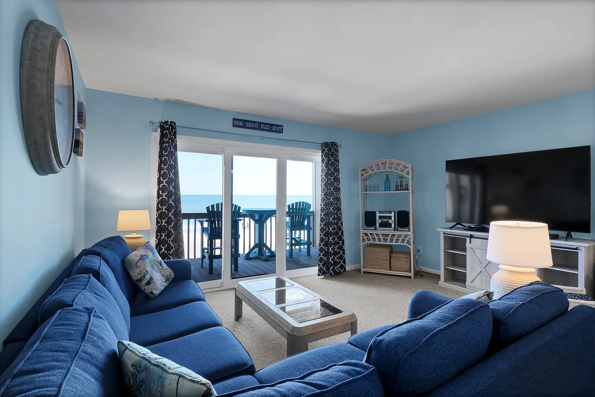 This interior image centers on a spacious blue sectional sofa facing large glass doors that open to a balcony with a clear ocean view. The decorative shelf features beach-themed accents, and the room is bathed in natural light, showcasing a casual and inviting atmosphere perfect for relaxing or socializing.