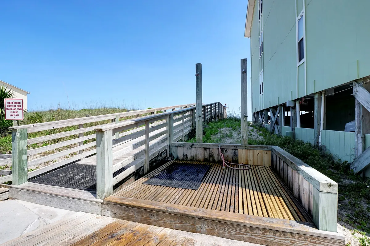 An inviting exterior shot of a wooden deck with a shower area and a sunny pathway leading directly to the beach. The area is bathed in warm light, emphasizing easy, secure, and direct access to the shore.