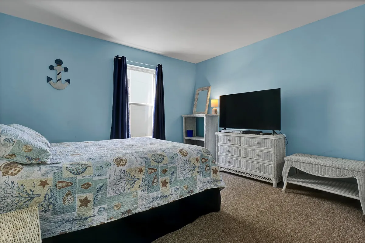 A tranquil master bedroom featuring a queen bed with ocean-themed bedding, dark curtains framing a window, white wicker furniture, and a television on a stand. The room is softly lit by natural light, creating an inviting retreat for relaxation or entertainment.