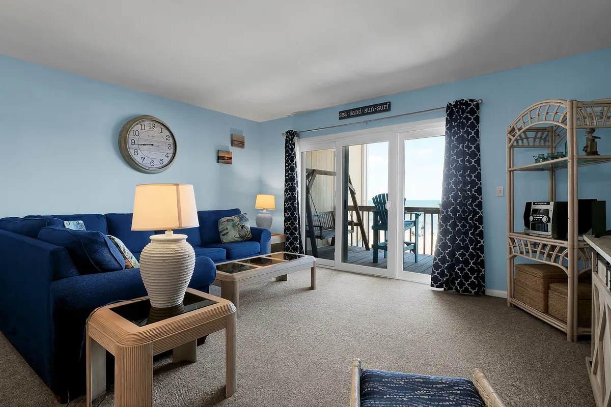 This image showcases a stylish living area with a blue sofa and coffee table, large windows that open onto a private balcony, and a decorative shelf. The space is calm and cheerful, with natural light pouring in and clear views of the ocean beyond the balcony doors.