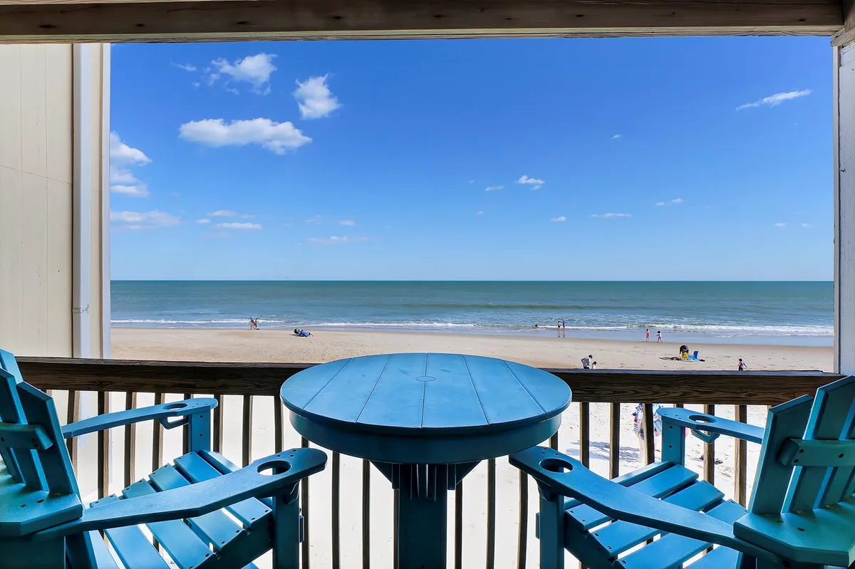 The image shows a serene first-floor oceanfront deck with bright blue chairs and a circular table, overlooking the sandy beach and sparkling waves. The scene exudes relaxation, with the promise of breathtaking sunset views, morning coffee with an ocean breeze, and the chance to spot dolphins playing in the distance.