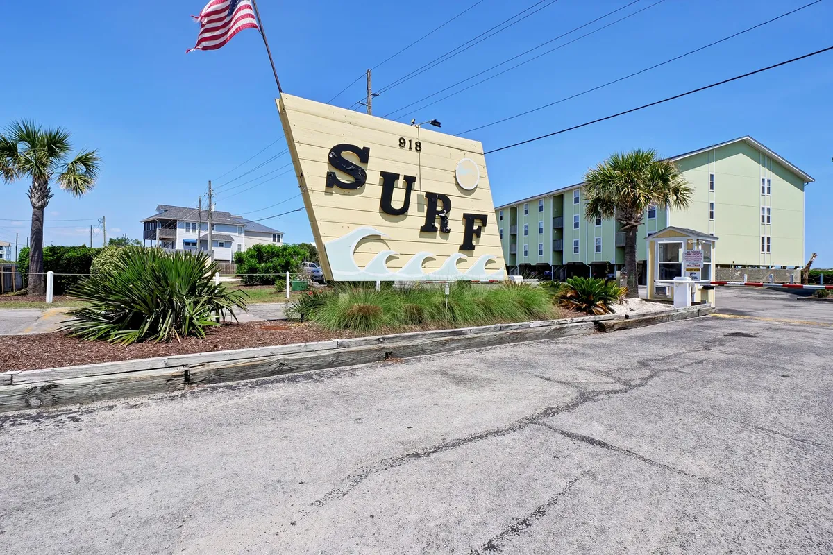 This exterior image features a large sign with the word 'SURF', palm trees, and nearby buildings. The relaxed scene captures the local vibe and hints at the proximity to the beach and Surf City attractions.