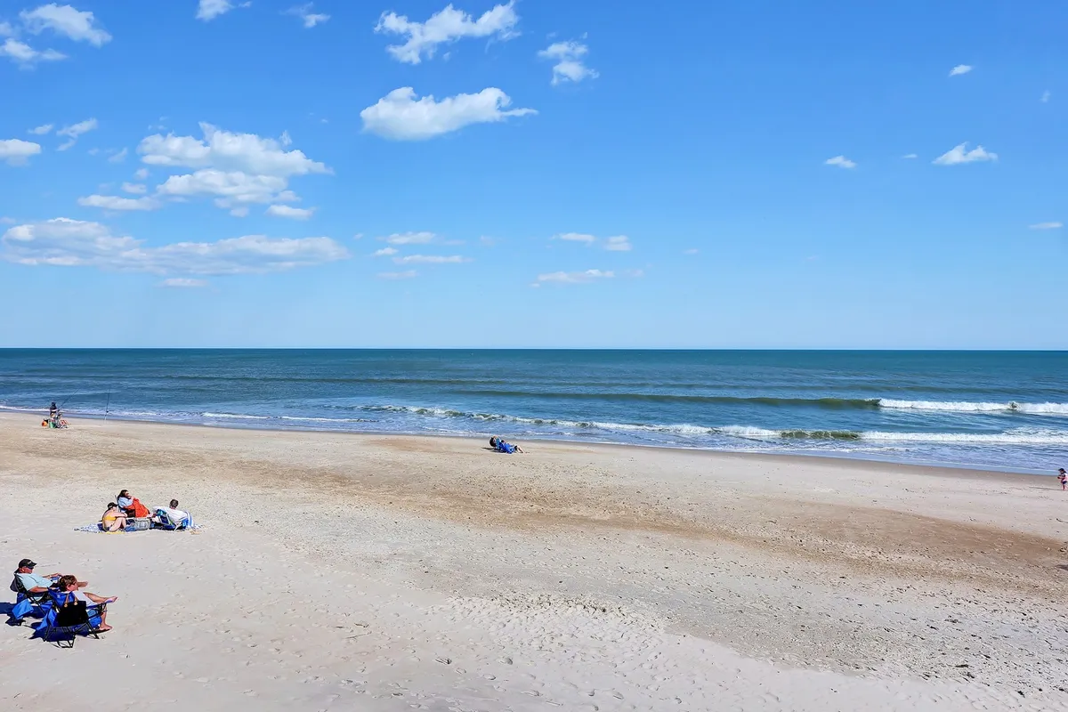 A wide beach scene with beachgoers lightly scattered along the sand, gentle waves rolling in, and the sun low on the horizon, painting the sky with warm colors. The horizon feels endless, enhancing the sense of escape and tranquility.