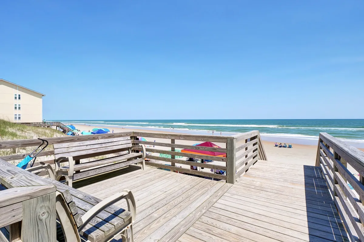 A wooden deck overlooking the ocean, with beach umbrellas and distant beachgoers visible. The view suggests easy transitions between sunbathing, beach games, and relaxed lounging, all within a scenic oceanfront setting.