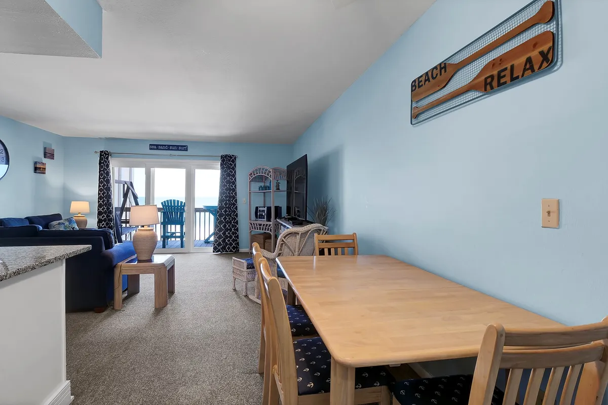 An open-plan scene highlights the blue-accented living area with a comfortable sofa, a dining table for shared meals, and direct access to the balcony. The ocean can be glimpsed through the windows, and the space is designed for both relaxation and togetherness.