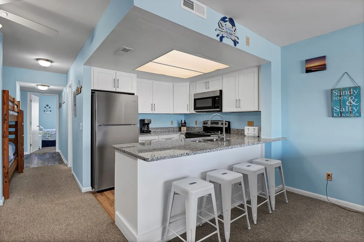 A cheerful kitchen scene featuring a granite-topped island with bar stools, stainless refrigerator and microwave, and decorative signs. The breakfast bar is ready for casual bites or morning coffee, while the arrangement encourages social connection.