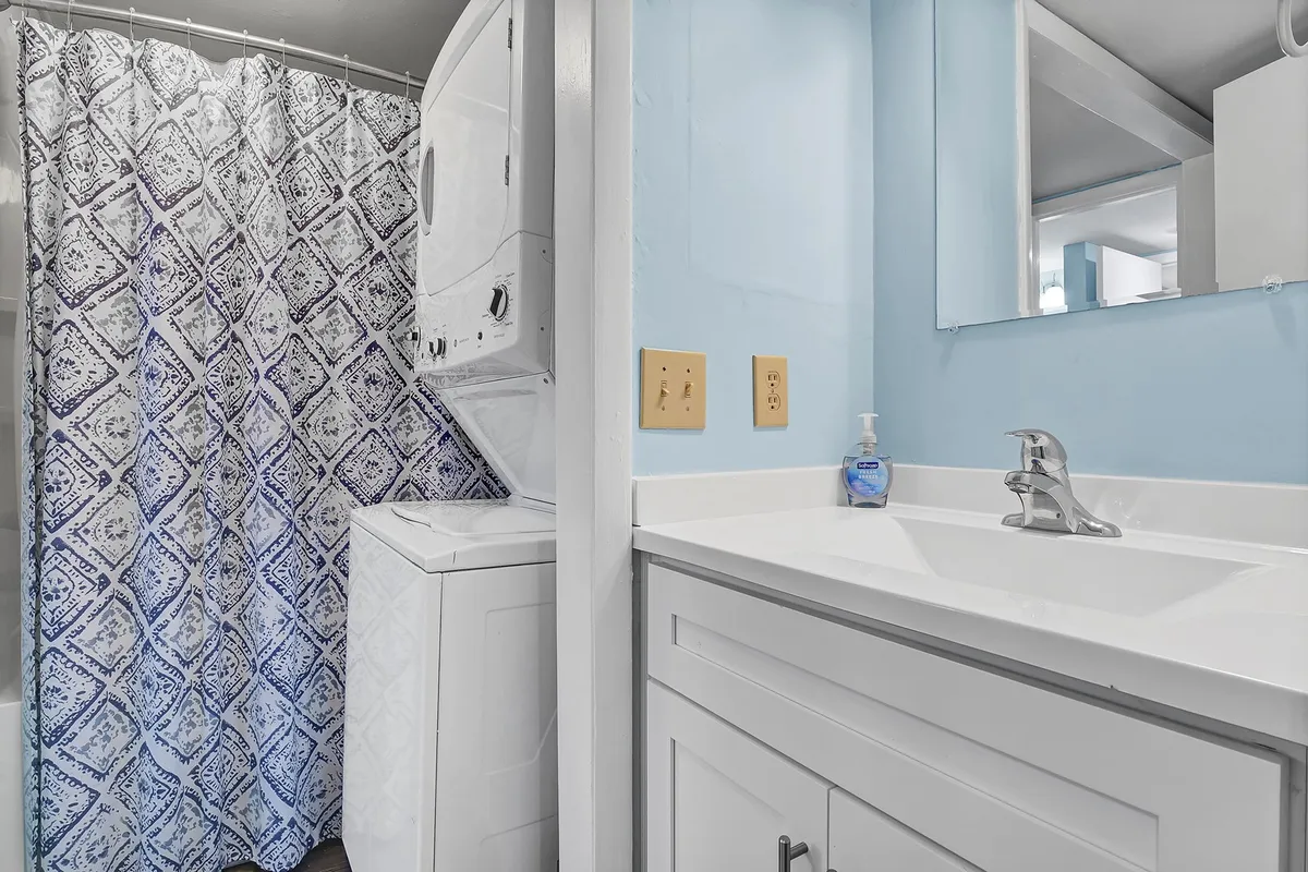 A practical interior scene shows a washing machine, shower curtain, and sink, alluding to the convenience of having laundry facilities and personal care amenities in the unit. The space is organized and user-friendly.