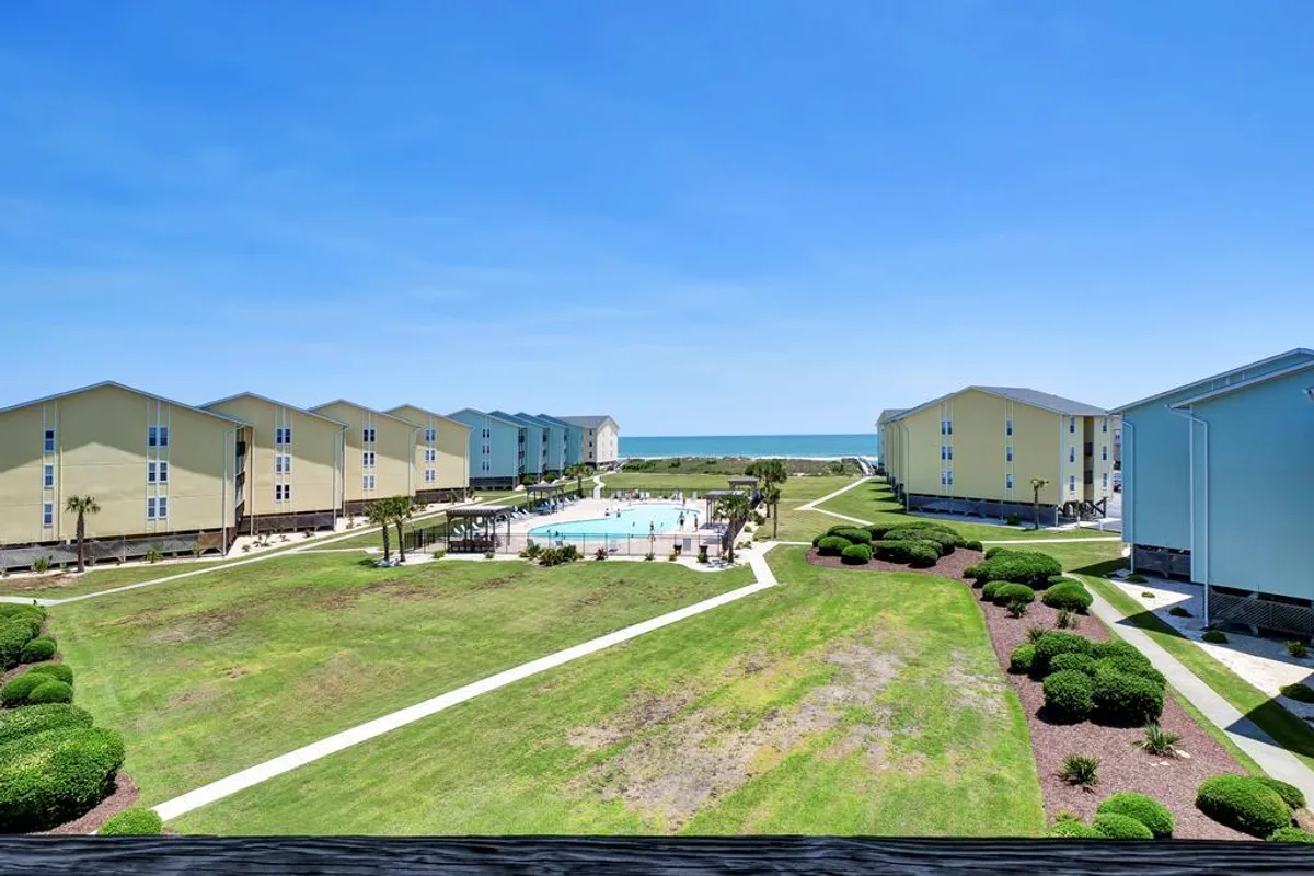 This image features the community swimming pool set against a green lawn, with a direct view of the beach and condominium buildings in the background. The pool area looks inviting with ample space for sunbathing and relaxing, while the proximity to the beach is clearly visible.