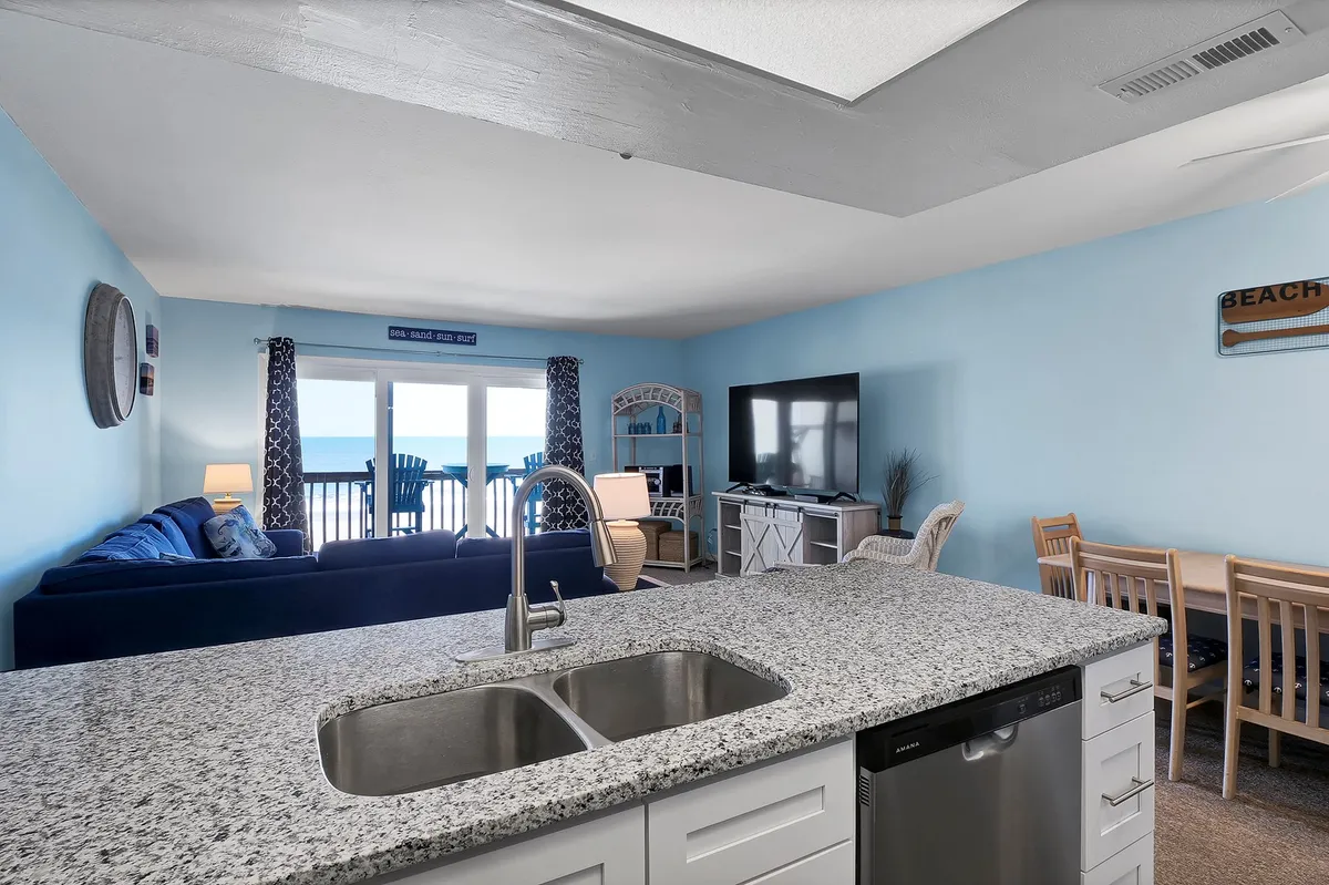 A bright, modern interior space showcasing a kitchen island, comfortable sofa, and large windows offering a clear ocean view. The open layout blends indoor comfort with outdoor scenery, hinting at shared meals and cozy relaxation.