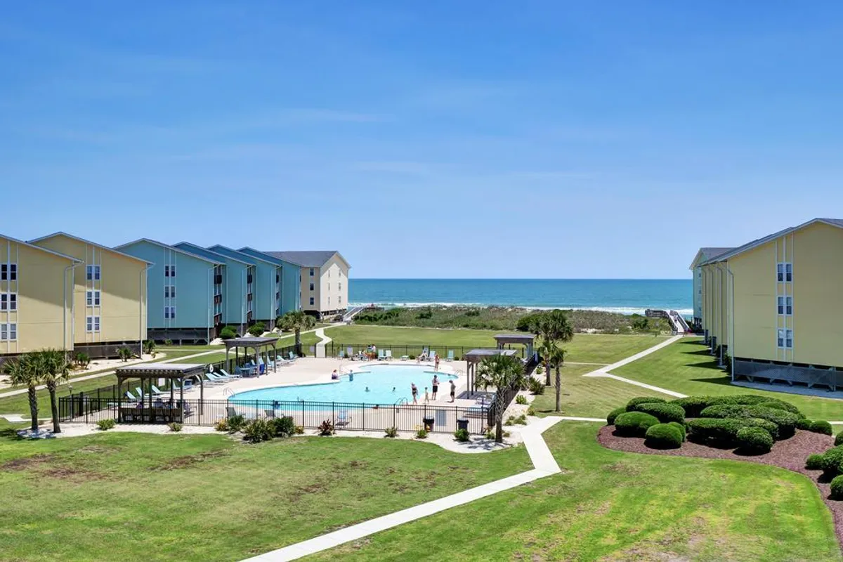 A vibrant outdoor scene featuring a swimming pool, landscaped gardens, colorful buildings, and a clear view of the beach in the background. People are shown swimming, sunbathing, and relaxing by the pool, reflecting the lively, cheerful atmosphere.