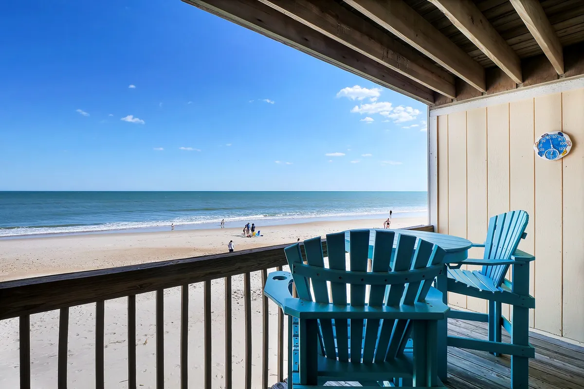 This image captures a tranquil beach scene, featuring adirondack chairs overlooking the ocean, sandy shoreline, and a calm wave pattern. The view suggests easy access to the Atlantic and highlights the natural beauty surrounding the property.