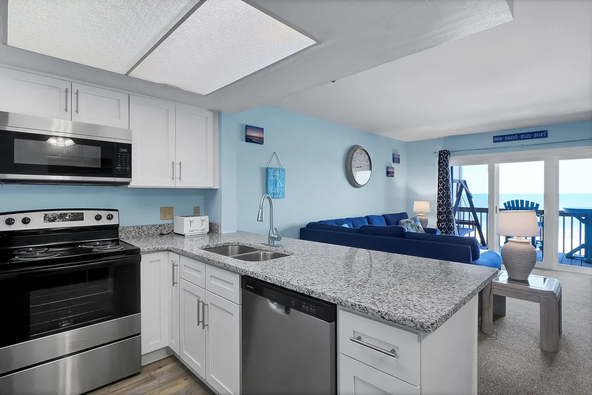 This image features a modern, open-concept kitchen with gleaming granite countertops, stainless steel appliances, and a blue couch that sits adjacent to the kitchen. Large windows offer an ocean view, blending the kitchen, dining, and living areas for a seamless, sociable atmosphere.